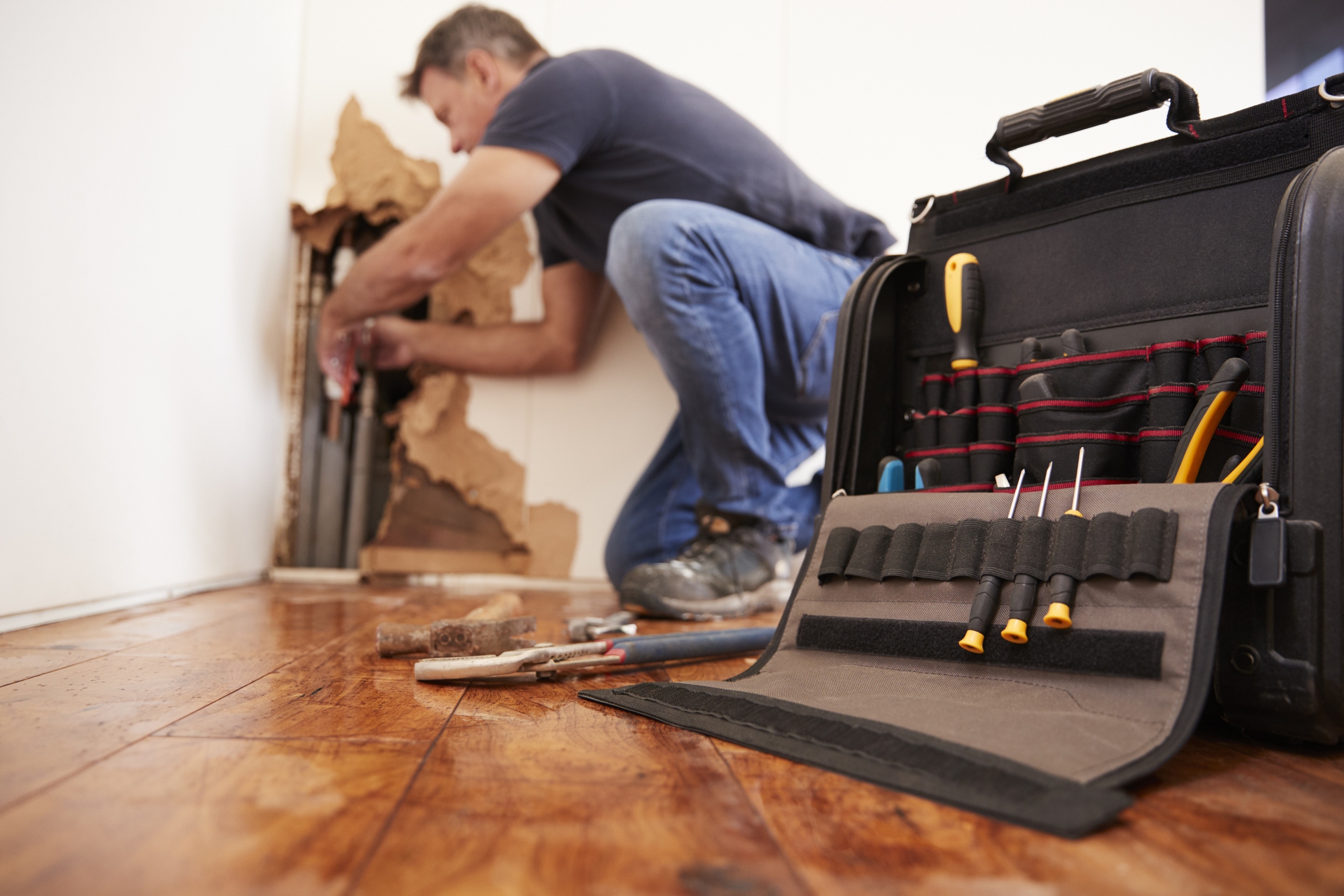 Technician performing slab leak detection through a hole in a home's wall, focus on tool bag in foreground.