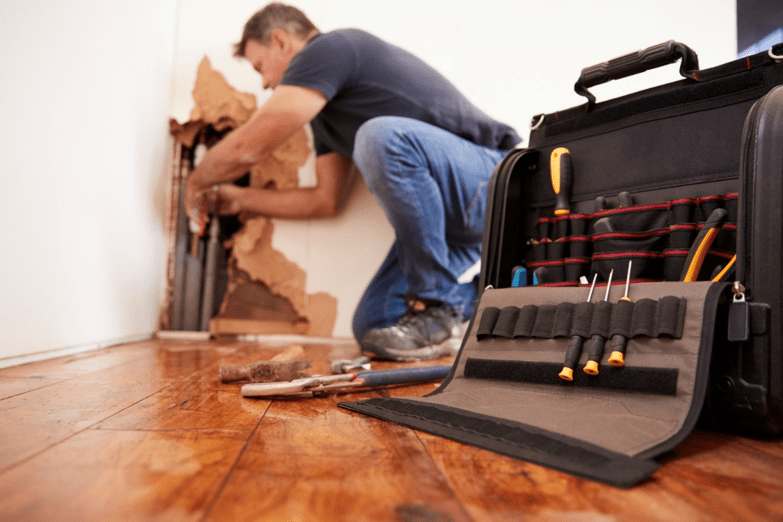 Plumber fixing a pipe in a home through a hole in the wall, with toolbox in foreground.